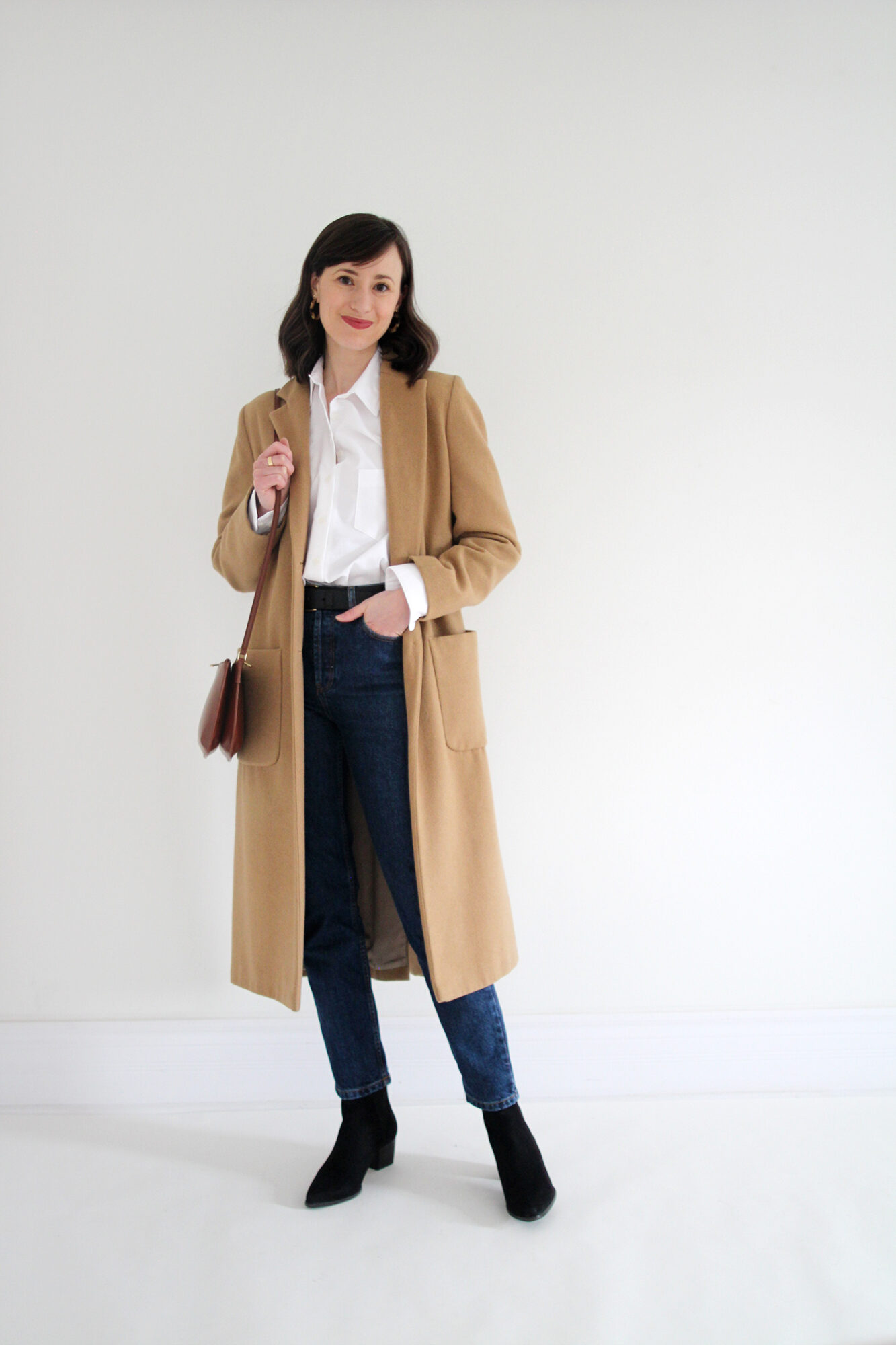 Woman wearing white button up, jeans, long camel coat, black boots, belt and brown leather bag.